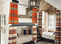 Beautifully styled cabin bunk room features a rustic built-in bunk bed complemented with striped curtains and drawers donning brown leather pulls. Sheepskin rugs sits sit on gray carpeting in front of the bunk bed and in front of a daybed hidden beneath a window and behind red, blue, and orange curtains.