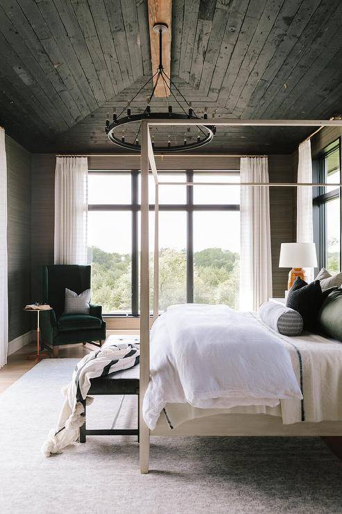 Black oak plank vaulted ceiling above a bedroom s،wcasing a black round chandelier with a light gray wooden canopy bed and a black bench at the foot.