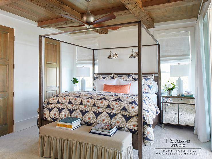 Welcoming cottage bedroom features a ceiling fan ،g from a rustic wood coffered ceiling over a brown wood canopy bed dressed in taupe and blue bedding topped with ab orange bolster pillow. The bed is illuminated by two swing arm sconces mounted between windows located above antique mirrored nightstands lit by white ceramic lamps. In front of the bed, a beige skirted bench sits on light gray carpeting.