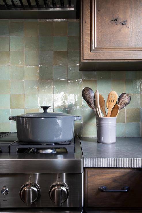 Kitchen features brown cabinets with concrete countertops and green glazed backsplash tiles.