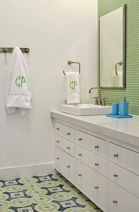 Blue and green kids' bathroom features accent wall clad in green glass grid tiles lined with white rippled cabinets with off-white counters topped with a rectangular vessel sink and a satin nickel faucet blue lacquer bath accents atop a green and blue geometric tiled floor.