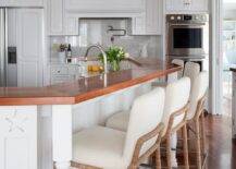 Ivory fabric and rope counter stools at a beadboard curved breakfast island in a cottage kitchen styled with a wood countertop complementing warm oak wood floors.
