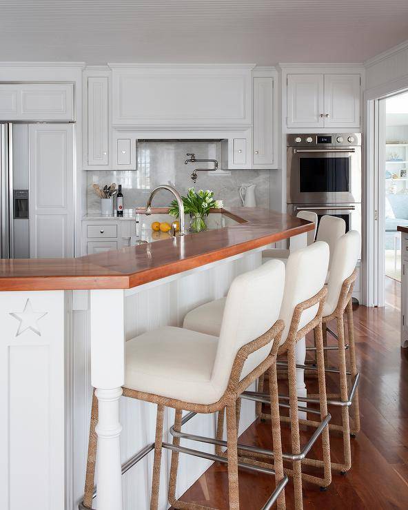 Ivory fabric and rope counter stools at a beadboard curved breakfast island in a cottage kitchen styled with a wood countertop complementing warm oak wood floors.
