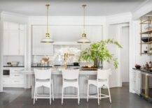 Spacious kitchen features modern white rattan stools at a light gray center island with brushed gold gooseneck faucet illuminated by white and gold lanterns, French brass bar shelves and white cabinets over glossy subway tiles.