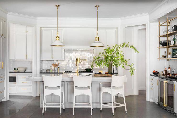 Spacious kitchen features modern white rattan stools at a light gray center island with brushed gold gooseneck faucet illuminated by white and gold lanterns, French brass bar shelves and white cabinets over glossy subway tiles.