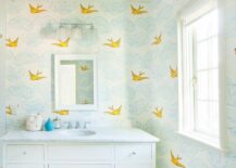 A whimsical and pleasant bathroom features blue and yellow Hygge & West Daydream Wallpaper with cloud like shapes and flying birds. A white framed bathroom window sill shines light on a single extra wide, white vanity cabinet with nickel knobs and honed white marble countertop. A polished nickel faucet sits under a white beveled mirror and 3 light frosted glass sconces.