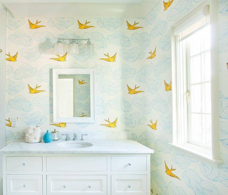 A whimsical and pleasant bathroom features blue and yellow Hygge & West Daydream Wallpaper with cloud like shapes and flying birds. A white framed bathroom window sill shines light on a single extra wide, white vanity cabinet with nickel knobs and honed white marble countertop. A polished nickel faucet sits under a white beveled mirror and 3 light frosted glass sconces.