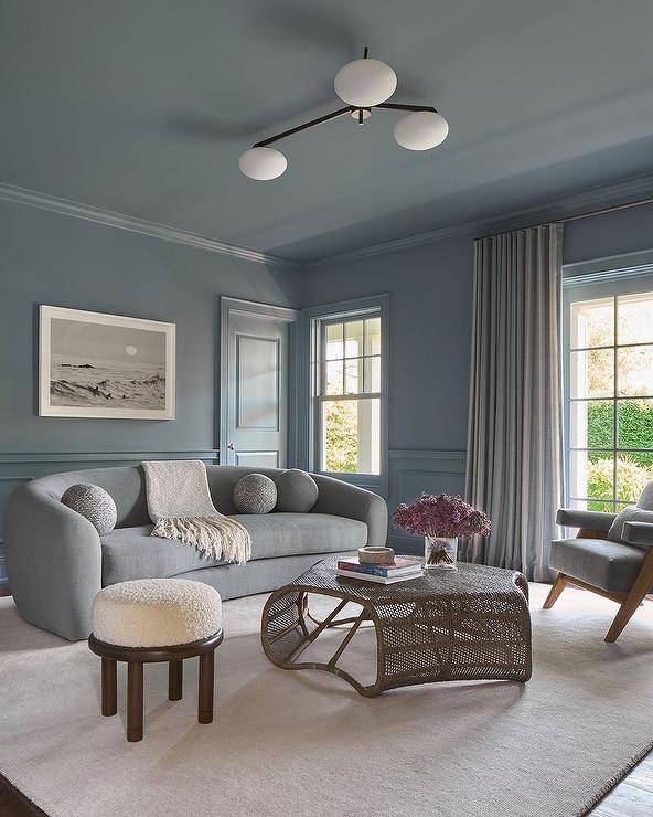 Living room features a curved gray sofa with a rattan coffee table, a gray accent chair under a window with gray curtains and a white stool atop a tan rug.