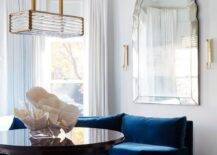 A unique brass and rippled glass linear chandelier lights a glossy round brown table placed on a blue rug in front of a curved dark blue velvet sofa. The sofa is positioned under a large Venetian mirror and in front of windows dressed in white curtains.