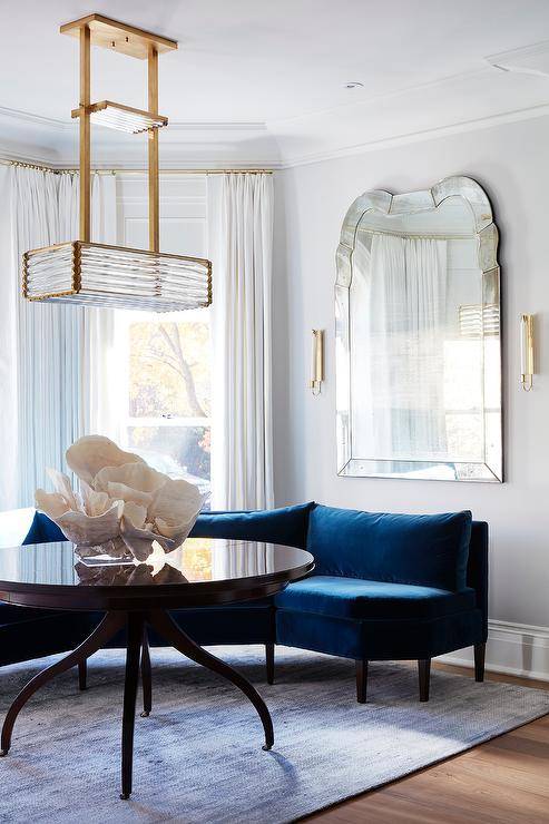 A unique br، and rippled gl، linear chandelier lights a glossy round brown table placed on a blue rug in front of a curved dark blue velvet sofa. The sofa is positioned under a large Venetian mirror and in front of windows dressed in white curtains.