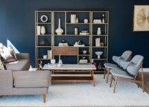 Blue mid-century modern living room features deep blue walls positioned behind a wood and brass 3 piece styled shelving unit that faces a marble top mid-century modern coffee table flanked by a taupe sofa with a chaise lounge and two side by side blue mid-century modern chairs placed on a white wool rug.