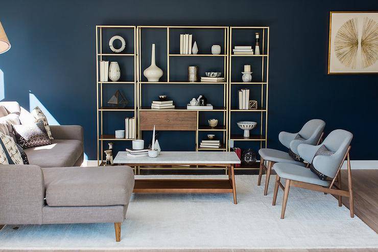 Blue mid-century modern living room features deep blue walls positioned behind a wood and brass 3 piece styled shelving unit that faces a marble top mid-century modern coffee table flanked by a taupe sofa with a chaise lounge and two side by side blue mid-century modern chairs placed on a white wool rug.