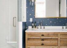 Kid's bathroom features a brass mirror mounted on blue and gold starburst wallpaper over a reclaimed wood dual washstand lit by a glass and brass sconce and a corner walk in shower.