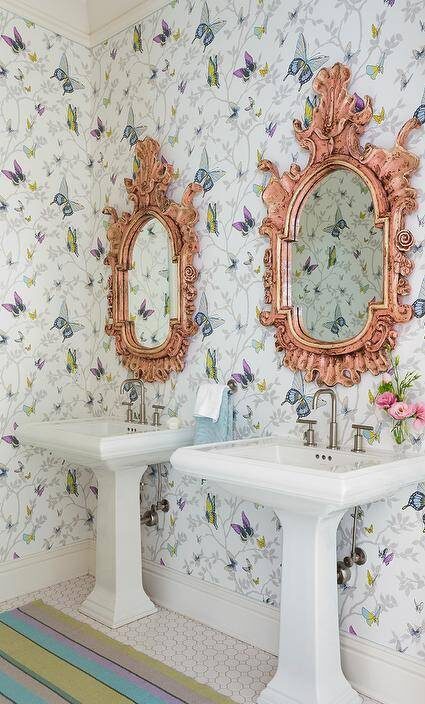 Colorful shared girls' bathroom with pink baroque mirrors mounted butterfly wallpaper above two white pedestal washstands, floor tiles are covered in a purple, blue and green striped rug.