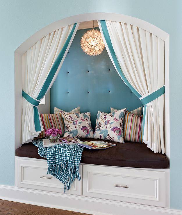 Framed by a wall painted in Sherwin Williams Waterscape, a white storage bench is topped with a brown cushion accented with blue and purple pillows placed against an blue tufted backsplash lit by a Capiz Flower Chandelier hung behind white and blue tie back curtains.