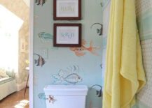 Stacked framed typography prints are hung on a wall covered in Nina Campbell Aquarium Wallpaper above a toilet positioned beside a wall accented with stripes of white hex and blue subway wall tiles fitted with brass towel hooks.