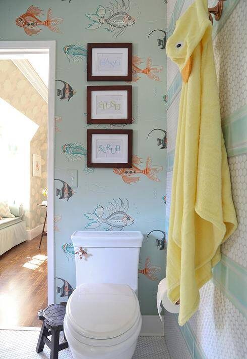 Stacked framed typography prints are hung on a wall covered in Nina Campbell Aquarium Wallpaper above a toilet positioned beside a wall accented with stripes of white hex and blue subway wall tiles fitted with brass towel hooks.