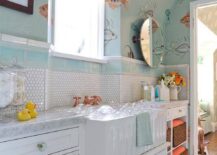 Gorgeous blue and white fish themed kids' bathroom boasts floor clad in white penny tiles placed beneath a white washstand fitted with glass knobs and a white apron sink paired with a brass faucet fixed in front of white hex backsplash tiles lined with blue subway border tiles mounted beneath a window framed by Nina Campbell Aquarium Wallpaper lit by a copper light pendant. Beside the washstand, wicker baskets stuff orange baskets fill shelves located under a carrera marble countertop placed under an oval pivot mirror position below a sloped ceiling,