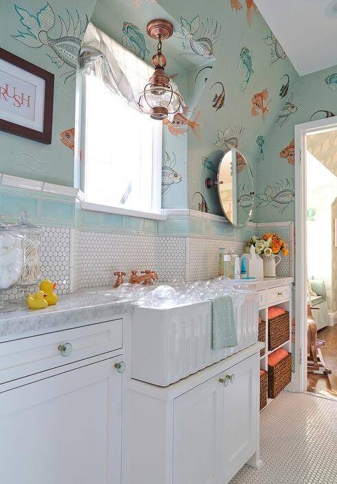 Gorgeous blue and white fish themed kids' bathroom boasts floor clad in white penny tiles placed beneath a white washstand fitted with glass knobs and a white apron sink paired with a brass faucet fixed in front of white hex backsplash tiles lined with blue subway border tiles mounted beneath a window framed by Nina Campbell Aquarium Wallpaper lit by a copper light pendant. Beside the washstand, wicker baskets stuff orange baskets fill shelves located under a carrera marble countertop placed under an oval pivot mirror position below a sloped ceiling,