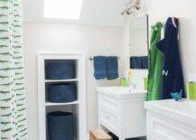 With natural light streaming in from a skylight, this well designed green and blue kids' bathroom is equipped with Serena & Lily Teak Step Stools placed on white hex floor tiles in front of white washstands adorning nickel pulls and polished nickel faucets placed beneath frameless vanity mirrors lit by 2 light nickel sconces. Towel racks separate the washstand as a Jonathan Adler Zebra Bath Mat is placed in front of a green lizard shower curtain facing niche shelves holding blue woven baskets.