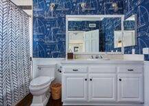 Clad in Ralph Lauren Chesapeake Wallpaper lined with a beadboard backsplash, this gorgeous blue and white kid's bathroom features a white washstand accented with oil rubbed bronze knobs and a white grid tile countertop. The countertop is fitted with an oval overmount sink paired with a polished nickel cross handle faucet mounted in front of a square tiled backsplash. A white framed mirror hangs above the washstand, while a toilet is positioned between the washstand and a drop-in tub covered with a Serena & Lily Feather Shower Curtain.