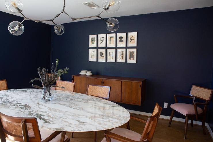 Oval Saarinen Dining Table joined with vintage cane chairs in a mid-century modern dining room finished with dark blue walls and an atom chandelier.