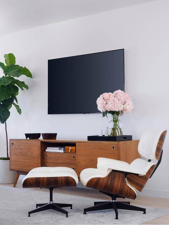 An Eames Lounger & Ottoman sits on a light gray rug in front of a mid-century modern TV console placed beside a fiddle leaf fig plant and under a flat panel TV.