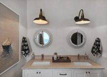 Shared boy's nautical bathroom is equipped with two large black vintage wall sconces mounted on gray walls over porthole mirrors fixed above a white dual vanity adorning oil rubbed bronze hardware and a gray stone countertop flanked by anchor towels and complementing a stunning paddle boat painting.
