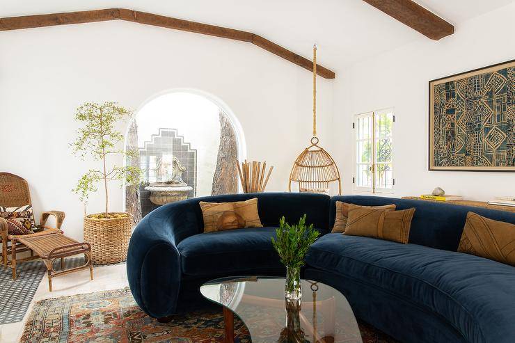 Vintage living room features curved blue velvet sofa with orange pillow on orange and blue vintage rug, vintage wood and glass coffee table, hanging rope rattan chair, wicker chair and ottoman over black and white mosaic floor tiles and potted olive tree that opens to an arched doorway that leads to a courtyard with a vintage lion water fountain.