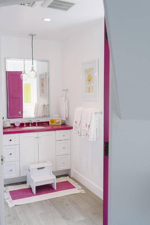 A hot pink door opens to a contemporary pink and white girl's bathroom featuring a white washstand finished with nickel hardware and a hot pink countertop holding an oval sink with a polished nickel faucet beneath a polished vanity mirror mounted on a white wall lit by light bulb pendant. A white step stool sits on a hot pink striped bath mat on gray wood-like floor tiles in front the washstand while a white and yellow framed print hangs above a polished nickel towel bar.