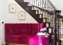 A contemporary foyer furnished with a stunning fuchsia velvet channel tufted sofa against a curved staircase wall with ornate iron spindles. A round acrylic accent table invites a luxurious touch to the curved sofa atop white and black checkered floors.