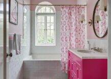 Lovely, pink girl's bathroom features a pink heart shaped bath rug placed on white marble honeycomb floor tiles in front of a hot pink washstand accented with glass knobs and a honed white marble countertop holding a round sink beneath a polished nickel cross handle faucet kit. The faucet is mounted in front of white subway backsplash tiles mounted beneath a round beaded mirror hung from a pink upper wall between glass and brass sconces. A subway tiled drop-in bathtub is covered with a pink medallion shower curtain hung in front of an arch window partially framed by white subway backsplash tiles.