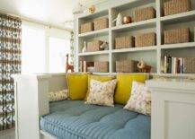 Gray built-in shelves lit by Aerin Charlton Sconces are fixed over a built-in reading nook topped with a blue cushion and yellow pillows.