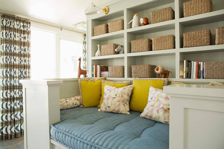 Gray built-in shelves lit by Aerin Charlton Sconces are fixed over a built-in reading nook topped with a blue cushion and yellow pillows.
