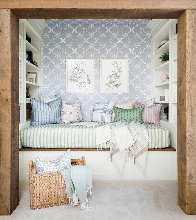 Rustic wood beams frame a reading nook boasting a built-in two-toned daybed flanked by white modular shelves and fixed against a wall covered in white and blue wallpaper finished with side-by-side art.