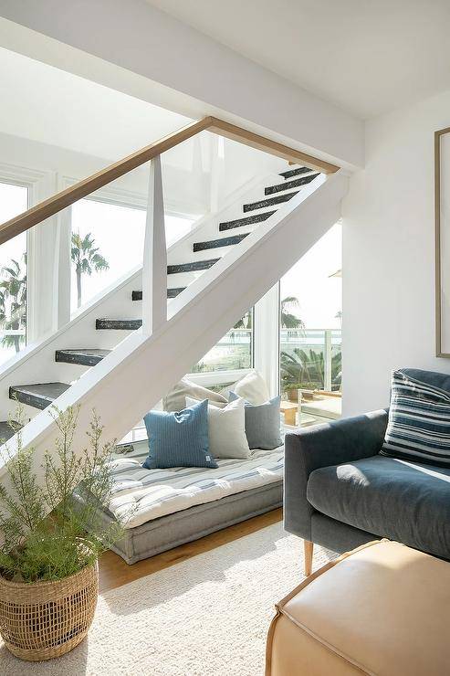 A small reading nook is located beneath a staircase and is accented with a rectangular gray floor cushion topped with a thin blue striped cushion and white and blue pillows.
