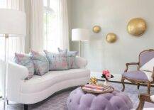 Arched windows framed by a light gray wall and covered in white curtains are positioned behind a white curved sofa topped with pink and blue pillows and placed on a purple chain link rug between gl، and nickel floor lamps. White and purple French chairs sit facing a purple velvet tufted ottoman coffee table placed in front of the sofa.