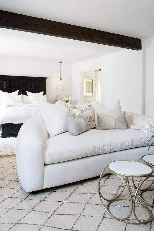 Under a dark stained wood ceiling beam, a white blush curved sofa sits on a Moroccan style wood rug in a bedroom sitting area featuring a white and gold accent table. Behind the sofa, on a white wool rug, a Restoration Hardware 17th C. Castelló Bed With Footboard is lit by a blow glass pendant hung over a nightstand.