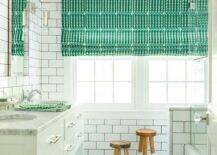 White and green shared boys' bathroom boasts Serena & Lily Dip Dyed Stools placed on Carrera marble hex floor tiles under a window dressed in a green and white geometric roman shade lit by a nickel sconce mounted on white subway wall tiles with gray grout fixed beside a chrome framed vanity mirror. The mirror is located above a Restoration Hardware Hutton Double Washstand adorning polished nickel hardware and a Carrera marble countertop illuminated by a Marine Porthole Flush Mount.