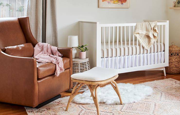 Vintage nursery design features a brown leather wingback glider with rattan stool and a white nursery crib with white and tan bedding atop a pink and gray nursery rug.