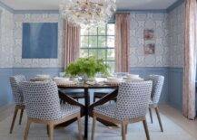 A glass bubbles chandelier hangs over a round black dining table matched with blue block print dining chairs placed on a light gray rug in a dining room clad in Galbraith & Paul Lotus wallpaper finished with blue crown moldings and blue wainscoting. Blue framed windows are dressed in pink curtains.
