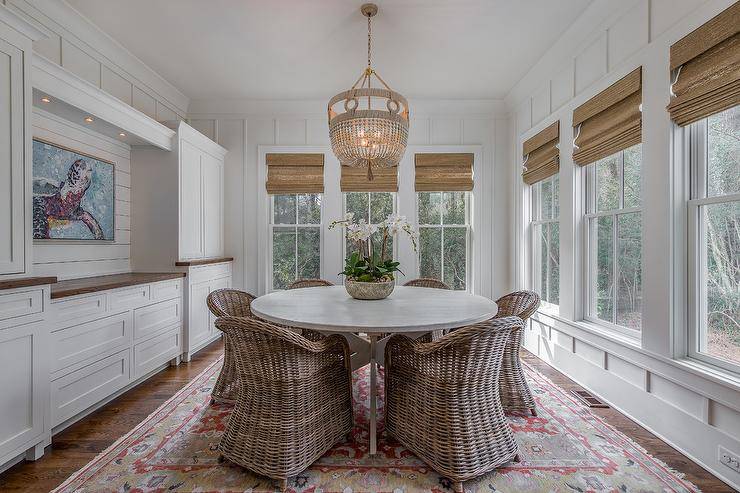 Cottage style dining room with a built-in sideboard accented with a wood top displaying an abstract wall art lit by a set of recessed lights. Ro Sham Beaux Frankie Malibu Chandelier decoratively hangs over a white round dining room table with wicker chairs over a gold and red Persian rug.