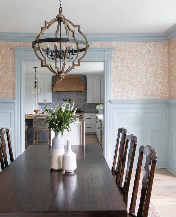 A wood and metal lantern hangs over a brown wood dining table surrounded by vintage wooden dining chairs. Walls half-covered in beige wallpaper are lined with blue crown moldings and finished with blue millwork.