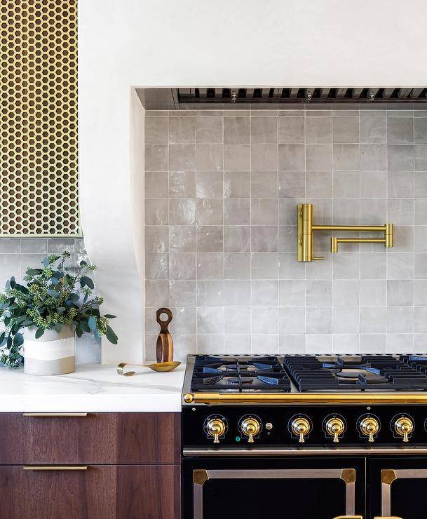 Modern kitchen features gray glazed tiles with a brass swing arm pot filler over a black French stove accented with brass hardware.