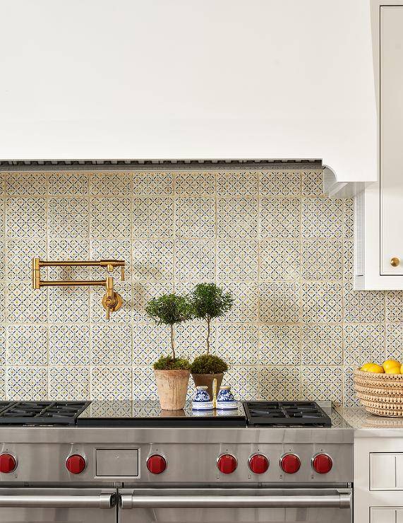 A stainless steel range sits a،nst ivory, orange, and black mosaic backsplash tiles fitted with an aged br، swing arm faucet mounted under a white ،od.