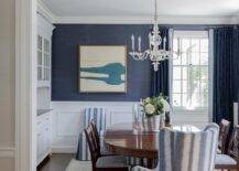 Wainscoting finishes dining room walls clad in blue grasscloth wallpaper. An ivory and blue abstract art piece hangs beside a window dressed in blue curtains and faces a French glass chandelier hung over an oval Chippendale dining table The dining table is matched with blue striped wingback head chairs and Chippendale dining chairs placed on an ivory and blue rug.