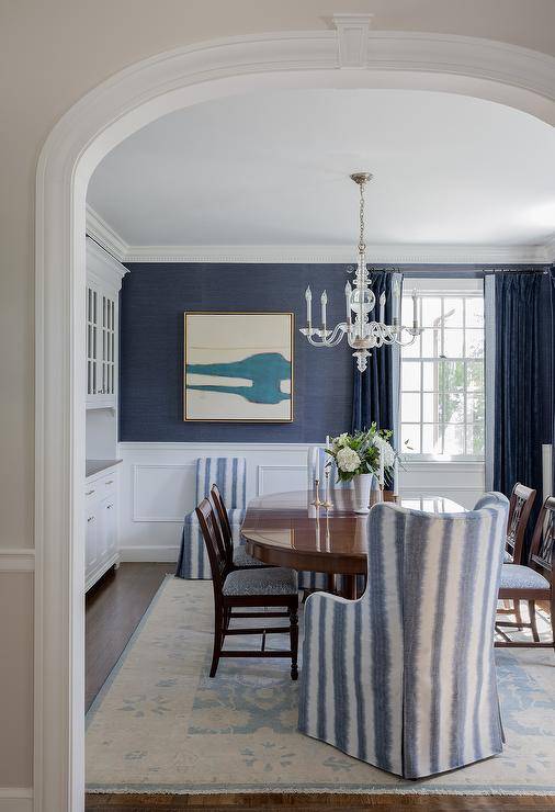 Wainscoting finishes dining room walls clad in blue grasscloth wallpaper. An ivory and blue abstract art piece hangs beside a window dressed in blue curtains and faces a French glass chandelier hung over an oval Chippendale dining table The dining table is matched with blue striped wingback head chairs and Chippendale dining chairs placed on an ivory and blue rug.