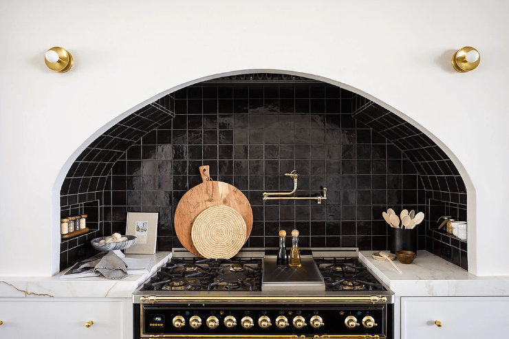 Kitchen features an arched cooking nook with glossy black grid tiles, a brass swing arm pot filler over a gold and black stove and white cabinet drawers with brass knobs and a marble countertop.