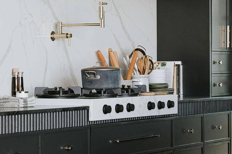 Kitchen features black cabinetry with black reeded trim and a polished brass swing arm pot filler on a marble slab backsplash.