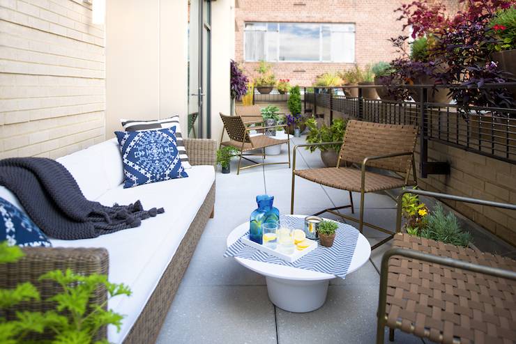 Private apartment terrace features outdoor woven sofa topped with white seat cushions, indigo blue pillows and a dark navy tasseled throw across from a pair of metal framed woven leather chairs alongside a modern round white coffee table.